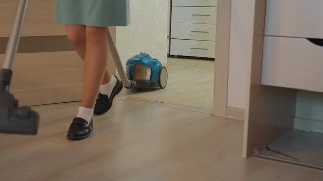 White schoolgirl vacuuming house hallway, pushing blue canister vacuum across hardwood floor, wearing white socks and black school shoes, neat bright interior, domestic chore and quiet morning routine