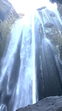 Low angle static shot of unidentifiable tourist silhouettes standing before thundering Gljufrabui waterfall cave