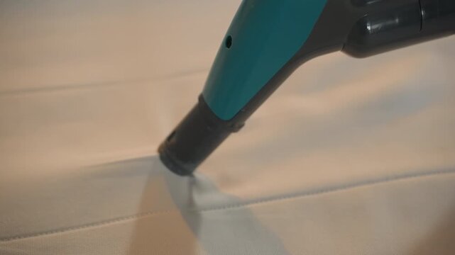 Closeup mattress cleaning with nozzle, handheld vacuum gliding over textured fabric, seams and fibers visible, gentle suction removing dust and light stains, warm indoor light, peaceful household