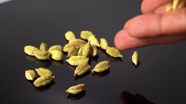 A close-up shot of green cardamom (Elettaria cardamomum) pods dropping from fingers onto a dark surface, highlighting the spice's texture and color