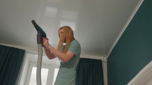 Blonde woman cleaning ceiling overhead, using steamer to remove dust from cornices while stretching and balancing on toes, focused expression. Teal bedroom, strong daylight through window, lowangle