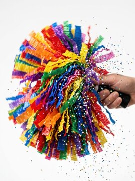 A young adult hand holds a colorful party popper with rainbow streamers exploding on white background.
