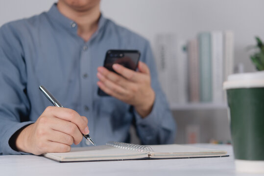 Businessman using smartphone and writing on notebook at desk for multitasking