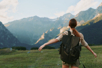 Obraz premium A woman hiker with a backpack stands in a meadow with arms outstretched toward distant mountains, capturing outdoor adventure, nature, and exploration in warm sunlight.