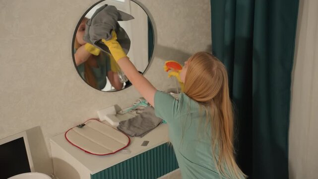 Young caucasian woman cleaning mirror in bedroom, wearing yellow rubber gloves and using microfiber cloth to wipe circular glass, reflection shows focused expression, bedside vanity with teal curtain