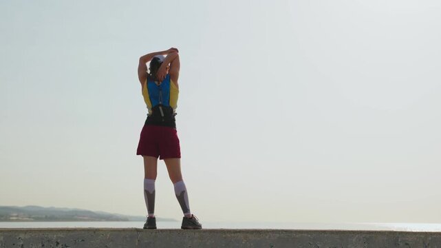 Woman stretches arms above head on sandy beach. Focused posture shows determination. Early morning light illuminates calm sea and hills. Close-up reveals tired yet resilient expression