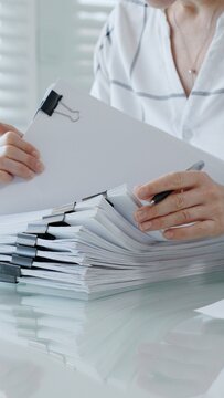 Person's hands meticulously reviewing and organizing extensive business documents and financial reports fastened with binder clips. Illustrating the demanding nature of administrative work