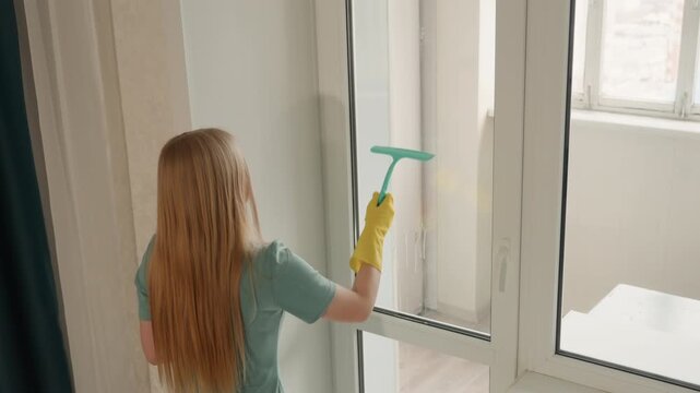 Caucasian woman squeegee cleaning tall window, wearing yellow gloves, systematic strokes and daylight, modern apartment balcony window, efficient chore, streakfree finish, focused movement