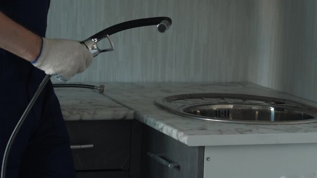 Plumber assembles a new kitchen faucet with a flexible spout before installing it into the sink. Experienced craftsman in protective gloves prepares water supply hoses for mounting.