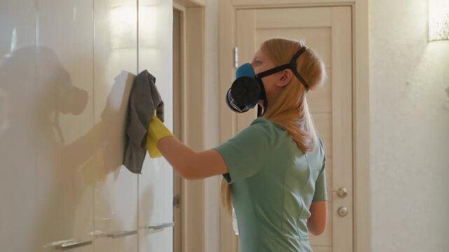 White woman wiping kitchen cabinets with cloth, wearing yellow gloves and mask uses green cleaner spray, steady routine, natural morning light, reflective glossy finish, calm domestic atmosphere