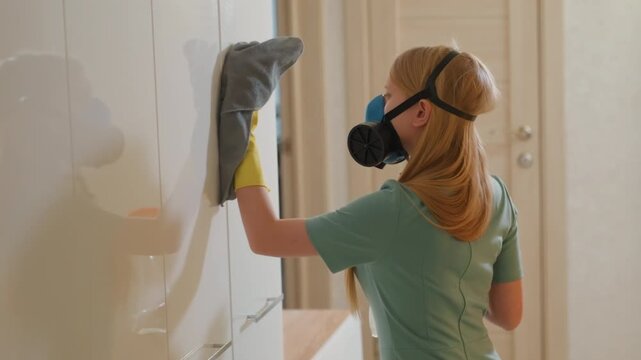 White woman wiping glossy cabinet doors with respirator and yellow gloves, using spray bottle and soft cloth to remove smudges and create streakless shine focused, careful strokes across reflective