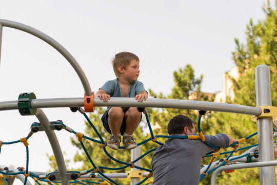 Young Boy Crouches on Playground Rope Net Above Father