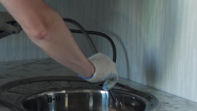 Plumber wearing protective gloves threads flexible hoses through the sink hole and installs the faucet. Professional craftsman carefully aligns and secures modern tap on the kitchen sink