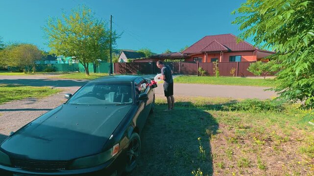 Man refueling from a gas can black car outdoors on sunny day near suburban house with red roof. Bright natural lighting, green trees, clear blue sky. Timelapse of the car filling up