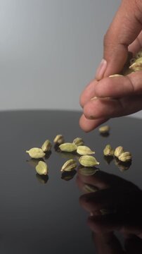 A close-up shot of an adult hand slowly releasing green cardamom pods onto a dark, reflective surface, creating a small pile of the aromatic spice