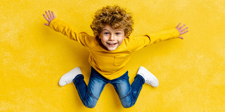 A young boy with curly hair is jumping with joy, wearing a yellow shirt and blue jeans, with arms and legs spread wide, against a yellow background.
