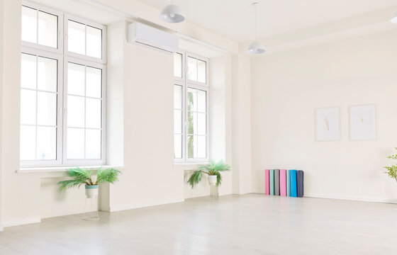 Minimalist yoga studio interior with windows, mats. Spacious white room with wooden floor, potted plants, and sunlight. Framed art and pendant lights. Peaceful space for mindful movement.
