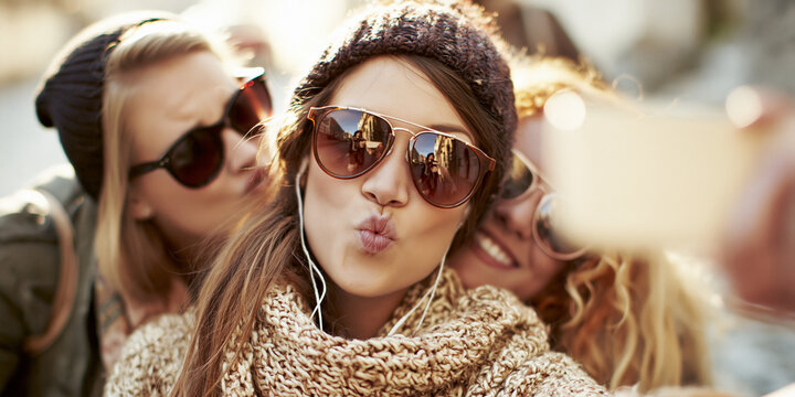 Three young women taking a selfie outdoors in winter, with one woman puckering her lips. They are all wearing winter clothing including beanies and sunglasses, and one woman is wearing earphones