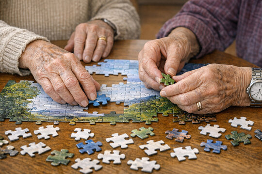 Two older individuals, possibly a couple, focused on completing a puzzle. They are holding and placing puzzle pieces together, engaged in friendly competition or cooperative play.