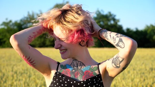 Portrait of happy punk girl with ear tunnels with blurred wheat field at background. Smiling hippie woman waving her pink hair and looking into camera at barley meadow. Concept of freedom. Slow mo