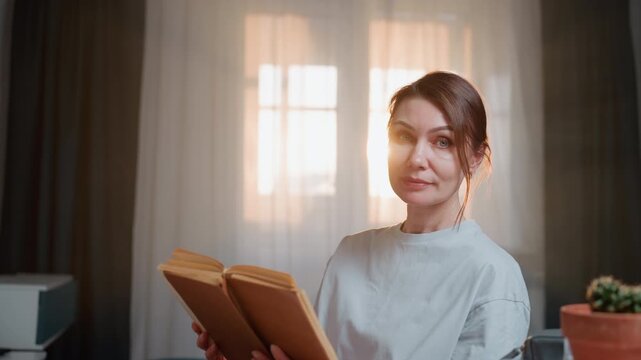 Woman reading peacefully. Lady immersed in book by sunlight. Contemplative woman with vintage notebook in cozy space. Serene female reader savoring solitary book time near luminous window