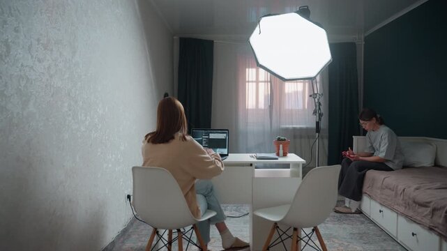 Home tutoring scene woman at desk with laptop while tutor on bed observes cozy bedroom studio lit by softbox grammar exercises on screen, notes and plant on table, focused study atmosphere and gentle