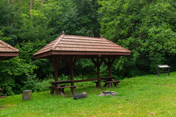 Obraz premium Open timber picnic shelter with benches and tables stands in a green woodland clearing beside a stone fire pit and information board. Quiet public recreation area designed for hiking breaks, family pi