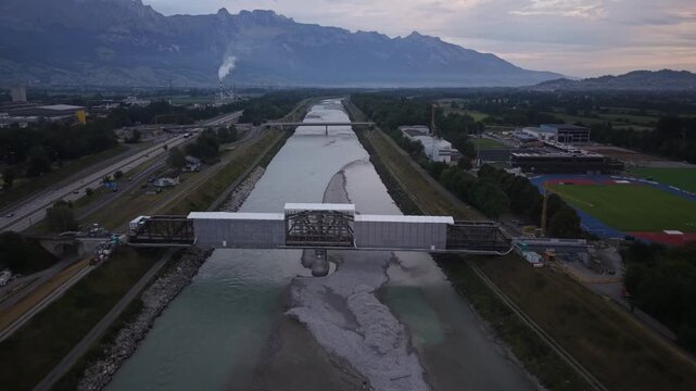 Forward drone flight toward a railway bridge undergoing renovation over the Rhine River, surrounded by extensive scaffolding and a temporary protective roof for construction work.