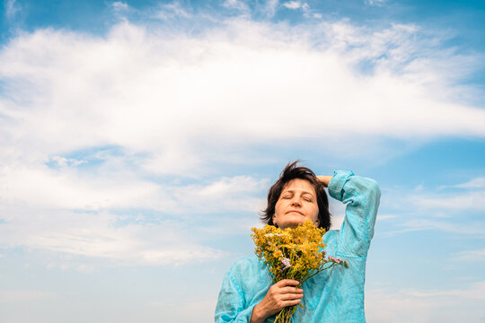 Woman stands outdoors, eyes closed, feeling the summer sun on her face and holding fresh bouquet of wildflowers