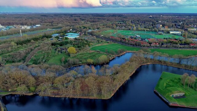 Aerial view video clip of Naarden star fort moat water, green earthwork bastions, surrounding forests, sports fields, residential neighborhood, and soft sunset clouds in North Holland, Netherlands
