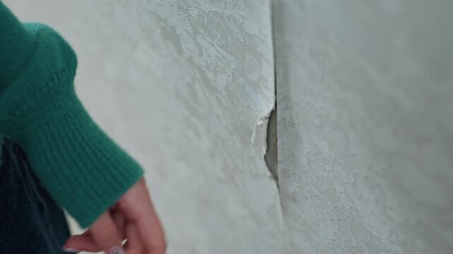 Closeup of peeling plaster and hairline crack in narrow hallway, woman in green sweater reaches with fingertip to assess damage, muted light, textured surface, contemplative mood, planning patch