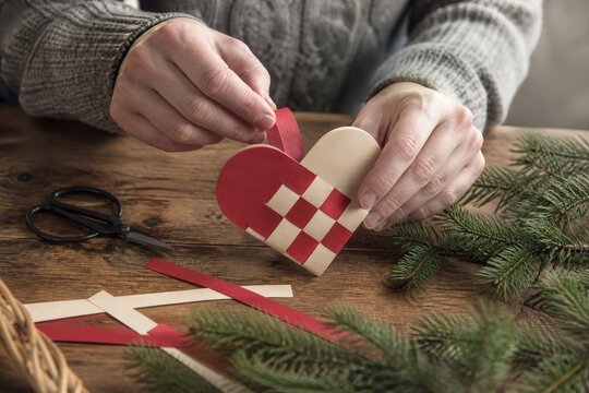 Hands weave a traditional Danish Christmas paper heart ornament on a rustic wooden table. This creative DIY activity embodies festive holiday traditions and artisanal craftsmanship.