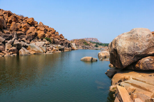 Scenic landscape of the Tungabhadra River flowing between massive ancient granite boulder hills and rock formations in Hampi, India. Historic nature, geological wonder and travel destination view.