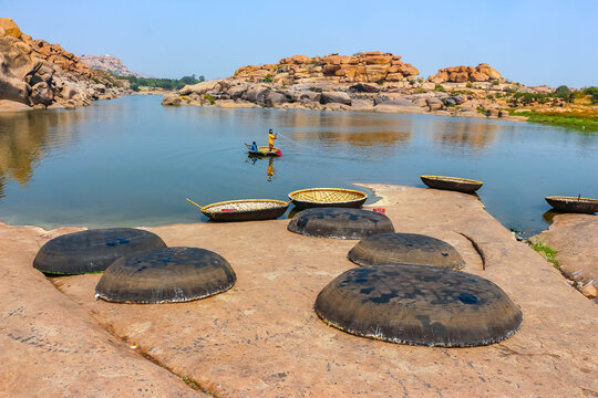 Traditional round wicker coracle boats upside down on the rocky shore of the Tungabhadra River with people floating on water in Hampi, India. Scenic landscape view, historic travel destination.