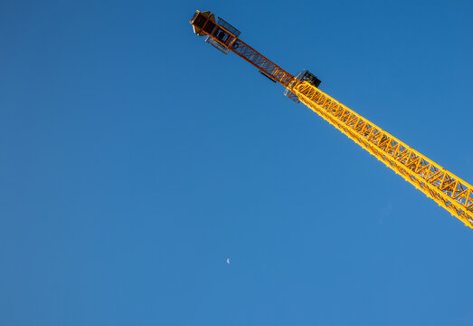 Yellow tower crane jib angled skyward against clear blue sky with moon visible