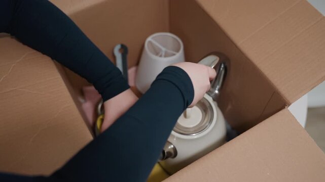 Caucasian hands unpacking kettle and teapot, arranges ceramic and metal pieces carefully inside box, uses dispenser and scissors to secure items, folds flaps and seals box, prepares kitchenware