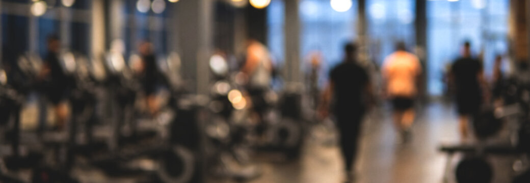 A blurred image of people exercising in a modern gym, featuring various workout equipment and bright lighting, conveying an energetic atmosphere.