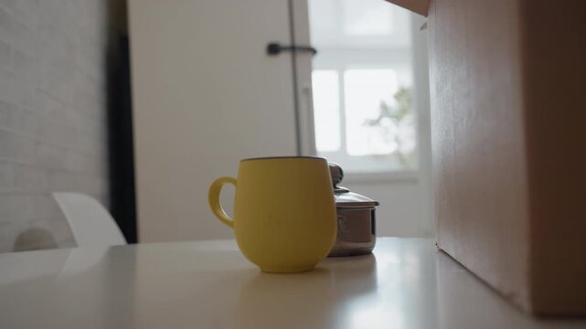 Yellow mug on countertop near box, daylight through window, kettle and teapot beside mug, cardboard box partially open, hand reaches momentarily then leaves empty table, calm domestic anticipation