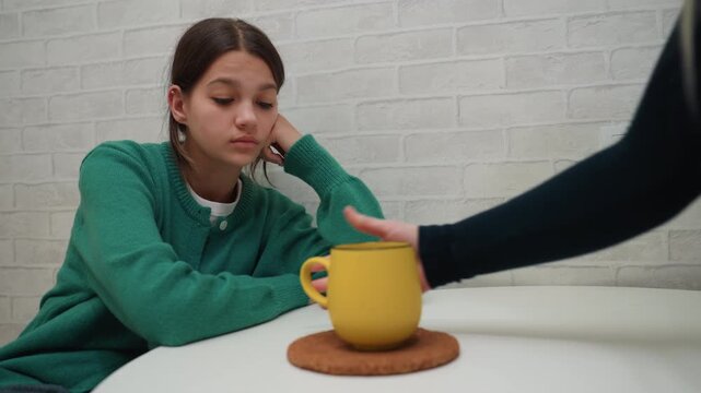 Girl at table with yellow mug, pensive new apartment kitchen scene teenager in green sweater leaning on white table against brick wall, hand places mug on coaster, quiet moving day mood with minimal