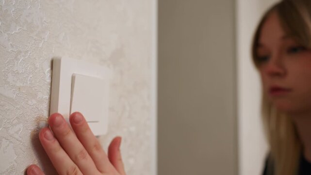 Young caucasian woman tests light switch in quiet new apartment hallway, introspective expression while unpacking boxes, hands touching textured wallpaper and plastic switch plate, soft natural
