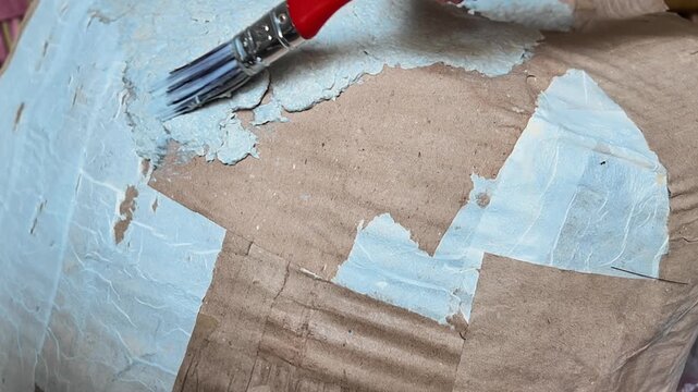 Handmade crafting process showing brush layering wet adhesive paste and paper sheets during DIY art project