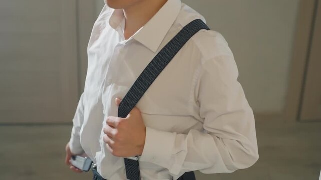 Asian man checking suspenders before leaving, confident posture and composed expression, hands smoothing shirt collar, crisp white collar and dark pinstripe trousers with clips, soft shadowing