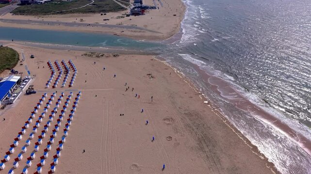 Aerial view video clip of Katwijk aan Zee beach resort, blue and red beach cabins, Oude Rijn river mouth, glistening North Sea waves, and coastal dunes in South Holland, Netherlands