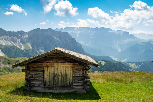 Rustic wooden mountain hut on Seceda alpine meadow overlooking Ortisei in Dolomites, Italy, with dramatic mountain peaks and valley in background under bright blue sky with clouds.