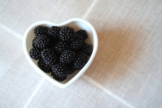 Heart-shaped bowl filled with ripe blackberries sits on a beige tablecloth. The arrangement highlights the fresh, nutritious appeal of the berries, enhancing their visual charm