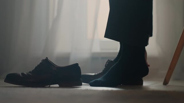 Closeup shoes and foot pulling sock, suitclad person sits on chair slipping into leather oxford, adjusting lace and ankle, soft morning light through sheer curtain, calm focused mood, preparing