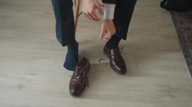 Asian man seated removing leather shoes, dark suit pants and white shirt cuffs, hands untying laces and placing polished brown shoes on light hardwood floor, quiet apartment evening, executive