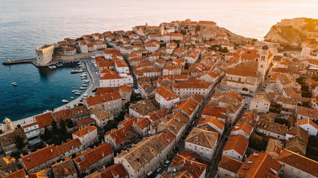 Aerial view of a coastal town with terracotta rooftops and blue sea under a warm sunset glow.