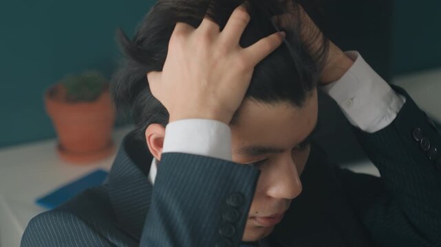 Young male businessman adjusting hair, quiet office desk with plant and laptop, careful grooming and nervous composure as he prepares for interview, subtle lighting highlights suit texture