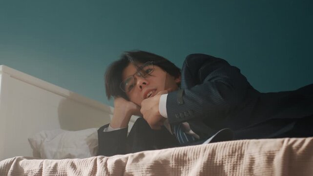 Contemplative individual resting quietly on bed. Asian male seated with calm and introspective expression. Pensive Asian man leans against headboard with soft light illuminating his thoughtful face